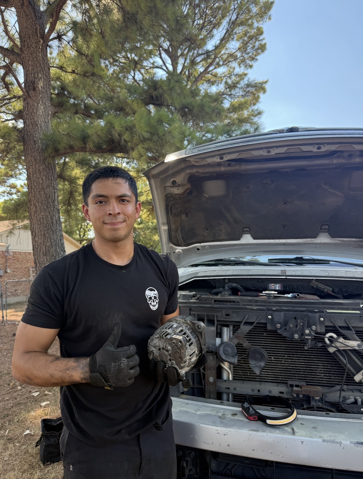 Alejandro, owner of AR Brothers Auto Repair, holding a replaced alternator next to a truck with the hood up
